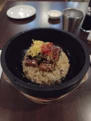 Unagi donburi on a cast-iron bowl atop a wooden platform, slightly soaked in teriyaki sauce, topped with sesame seed, ginger, and shredded egg. Empty sauce and sake cups are behind the bowl.