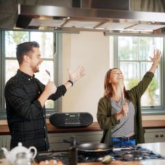 Two people in a kitchen singing onto their utensil on their right hand with their left arm raised. A superimposed boombox sits on a counter behind them.